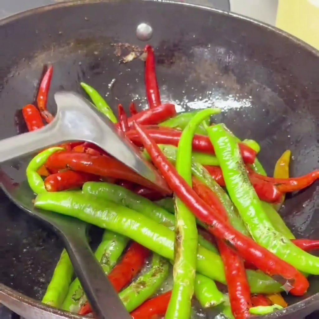 Blistered chili peppers being stir-fried with a spatula in a wok filled with shimmering spiced oil.