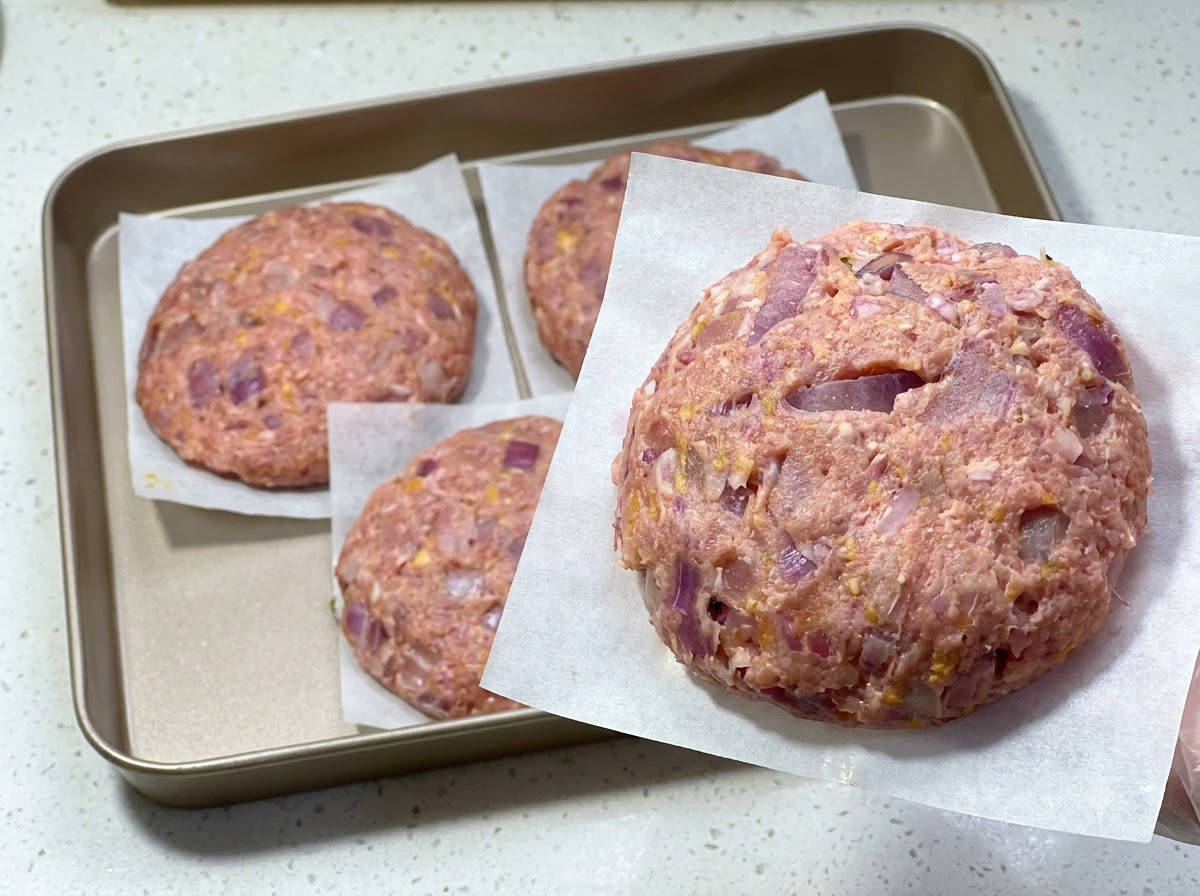 A freshly formed raw hamburger patty on parchment paper, with other patties set on a tray behind it.