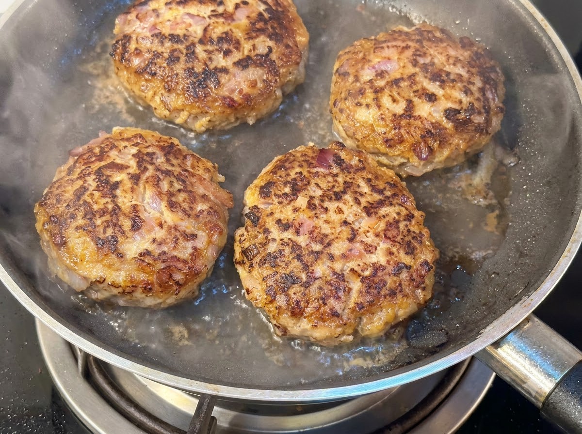 Four thick hamburger patties frying in a pan with water and steam rising underneath a lid.