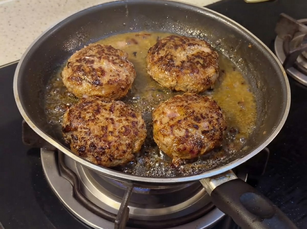 Hamburger patties cooking in a pan with liquid sauce ingredients starting to combine.