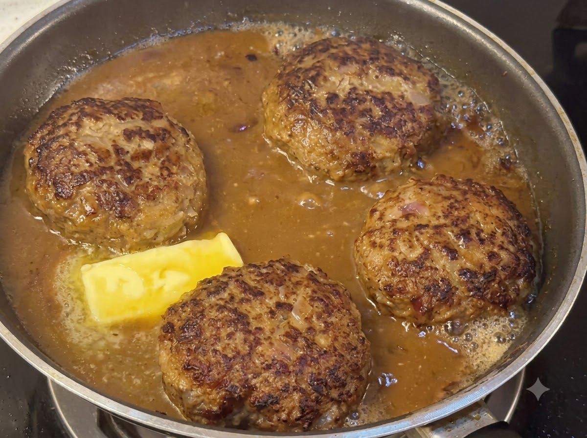 Hamburger patties simmering in a thick brown sauce alongside a melting piece of butter.