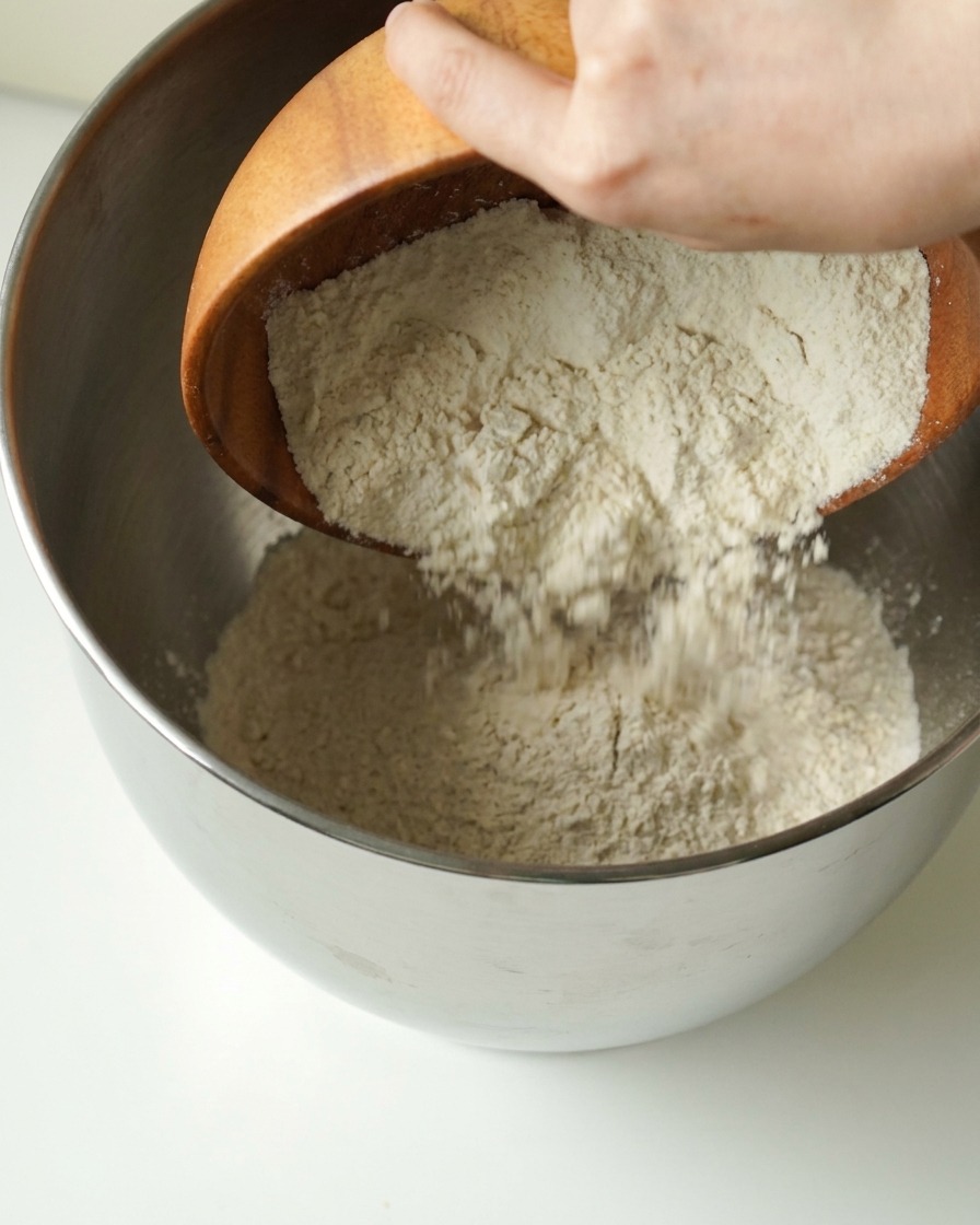 Flour being poured from a wooden bowl into a metal mixing bowl.