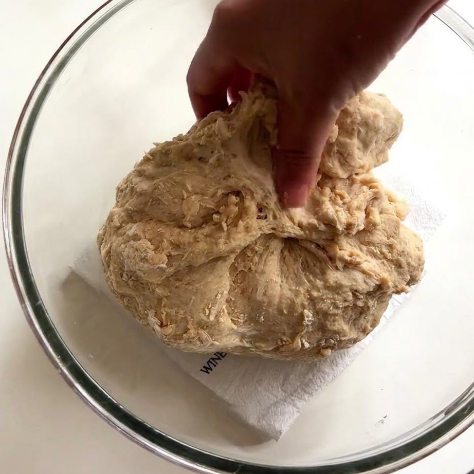 A persons hand using wet fingers to stretch and pull sticky bread dough inside a glass bowl.