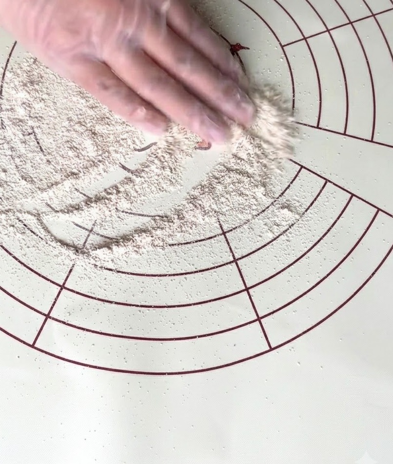 A gloved hand spreading whole wheat flour across a circular silicone kneading mat.