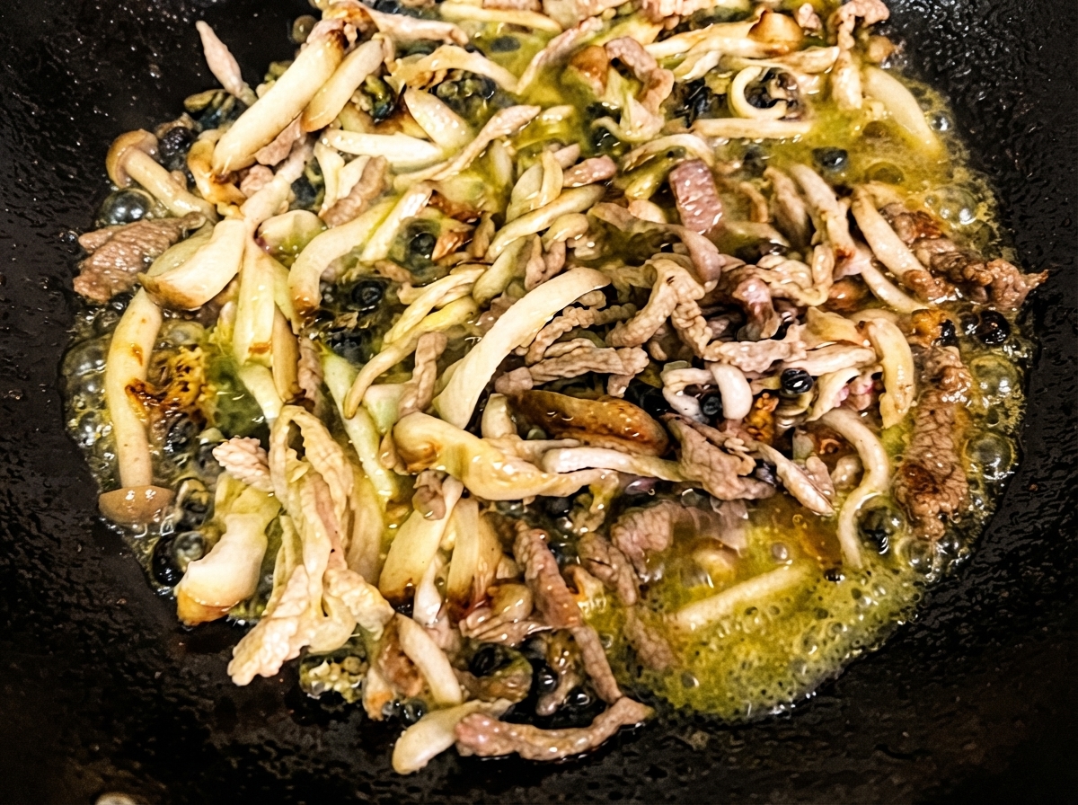 Shredded shiitake mushrooms, enoki mushrooms, and sea snails sautéing in hot oil inside a black wok.
