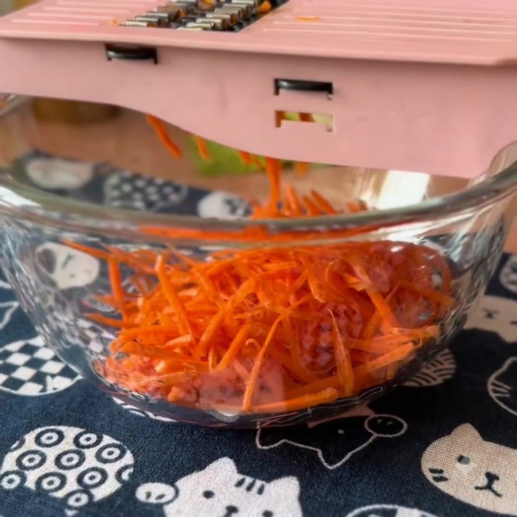 Fresh carrots and cucumbers being shredded with a pink mandoline slicer into a glass bowl.