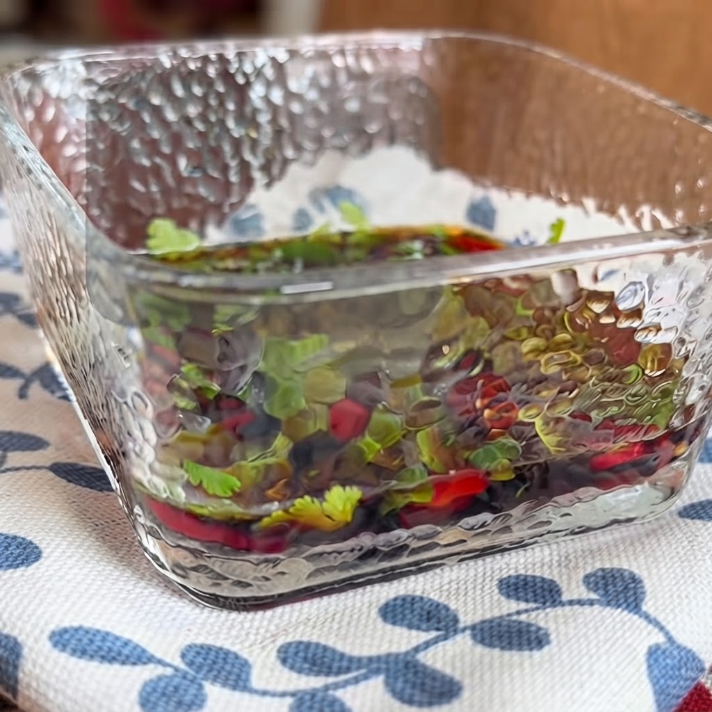 A close-up of a square glass dish filled with a savory dipping sauce containing herbs and chili peppers being mixed with chopsticks.