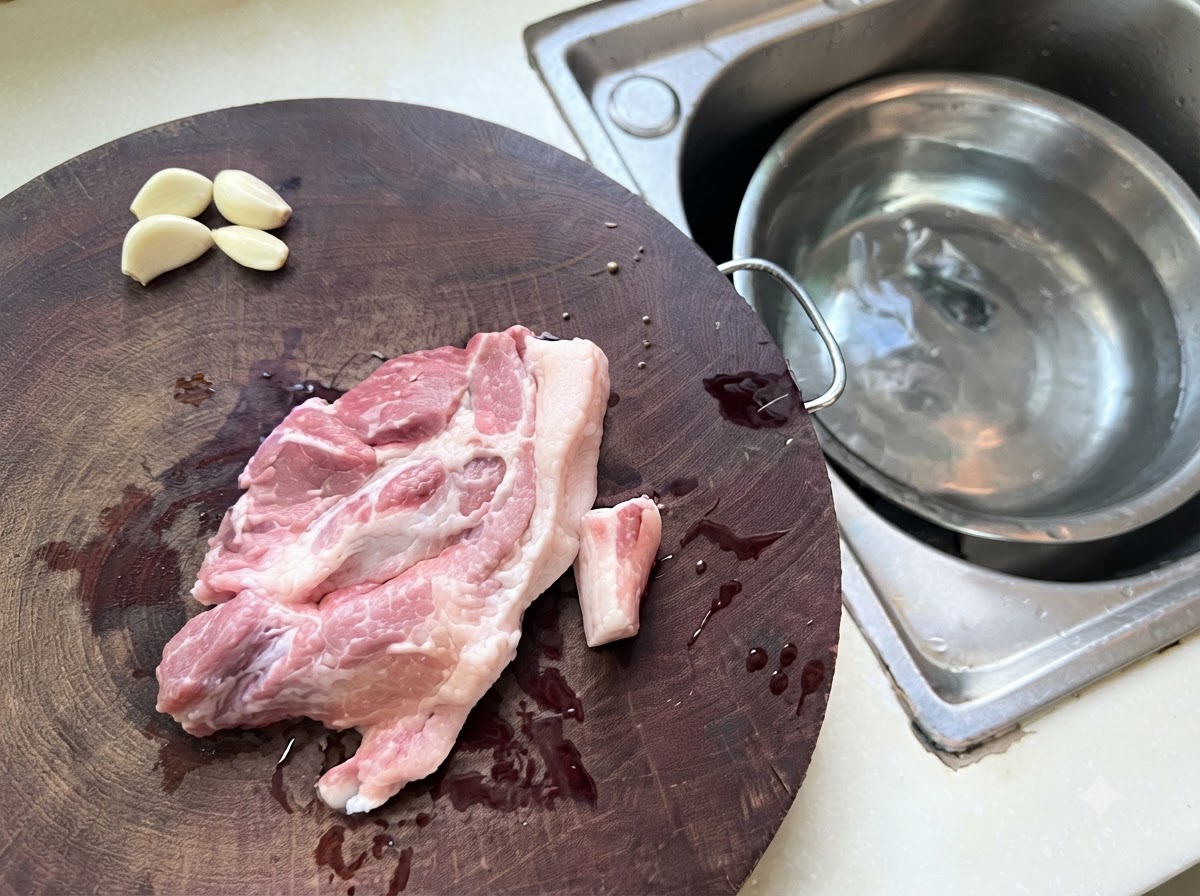 Minced pork and garlic pieces spread across a wooden cutting board next to a metal bowl of water.