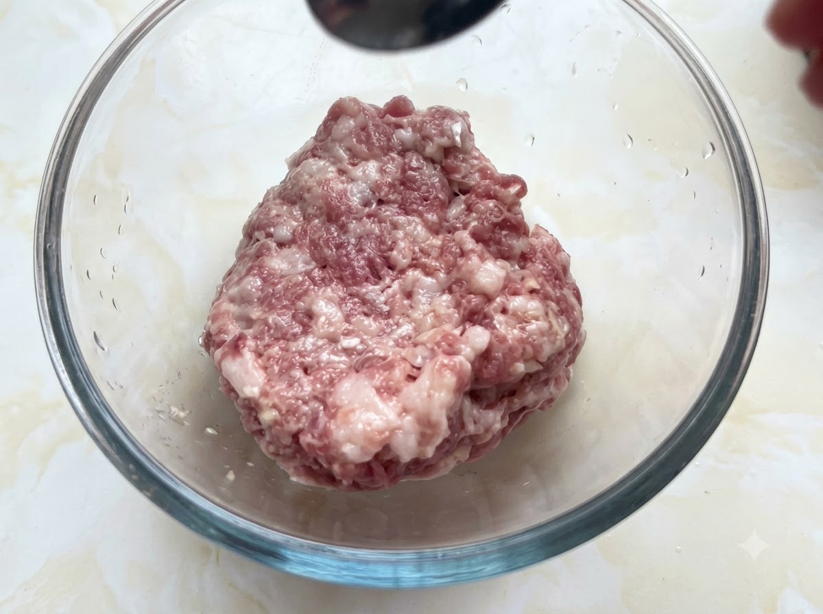 A glass bowl filled with minced pork receiving seasonings from a spoon held above.