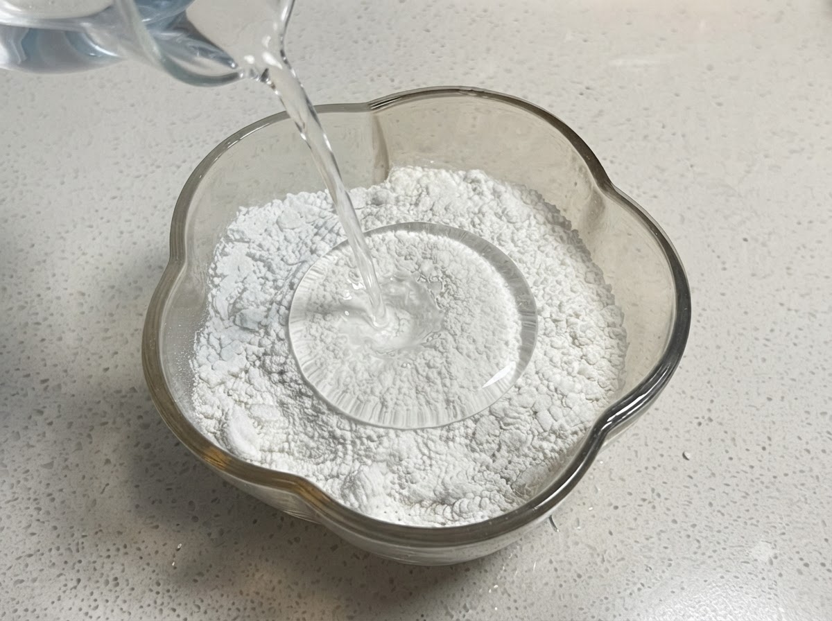 Water being poured from a measuring cup into a glass bowl containing a mix of flour and starch.
