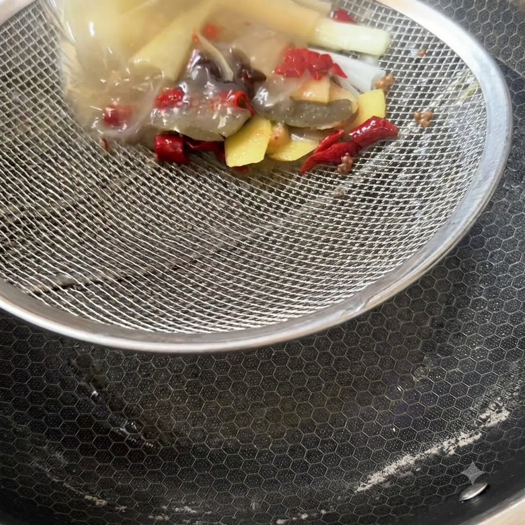 A strainer catching leftover spices, ginger slices, and chili dregs above an empty pan.