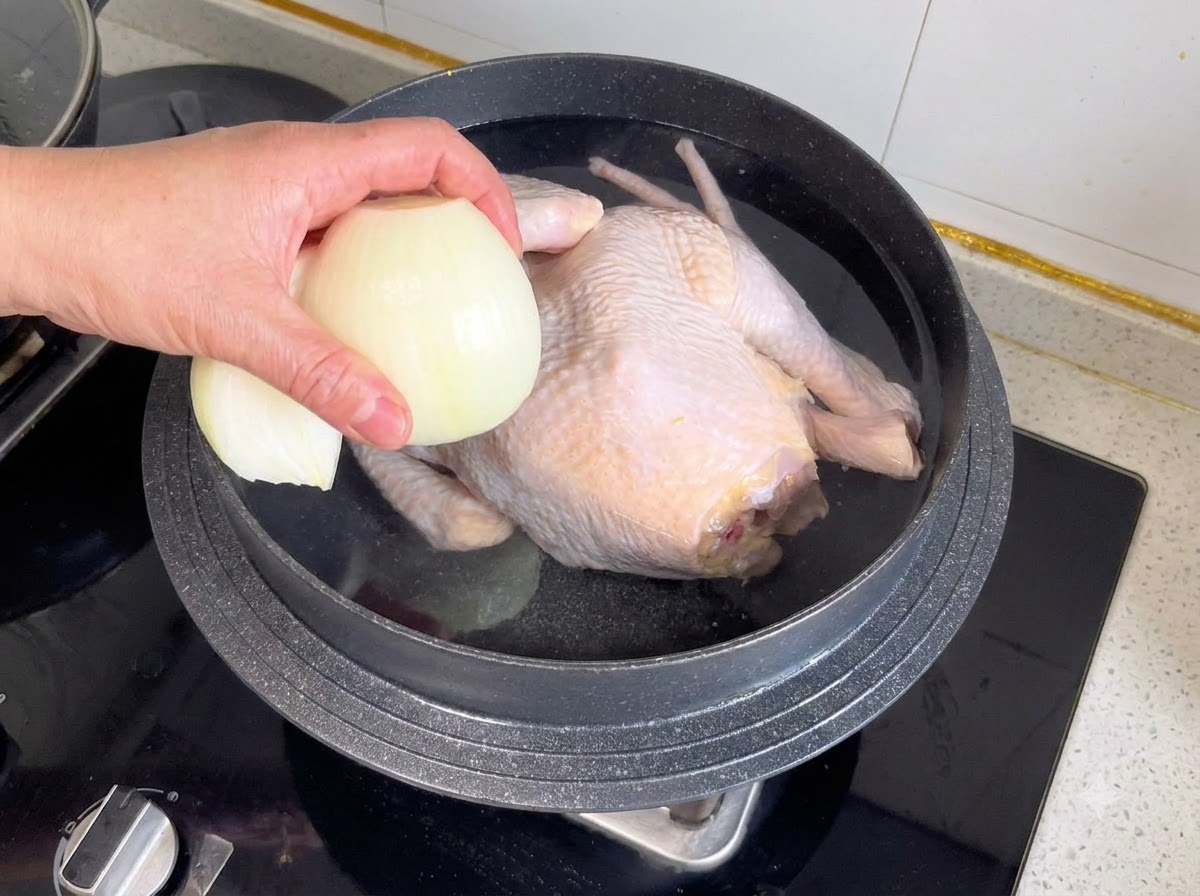 An onion half and other aromatics being dropped into a pot containing a whole chicken and water.