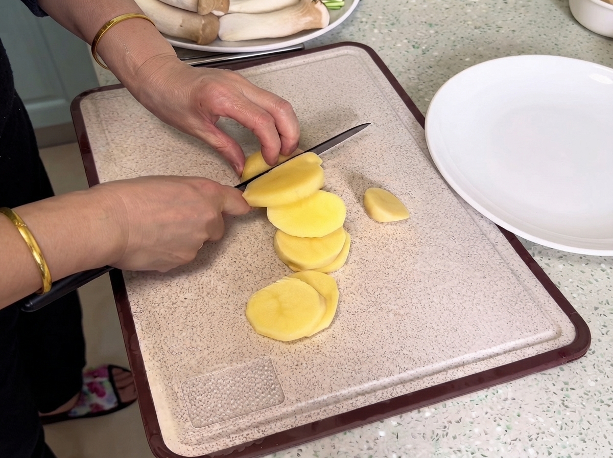 Potatoes being sliced with a knife on a cutting board next to a white plate.