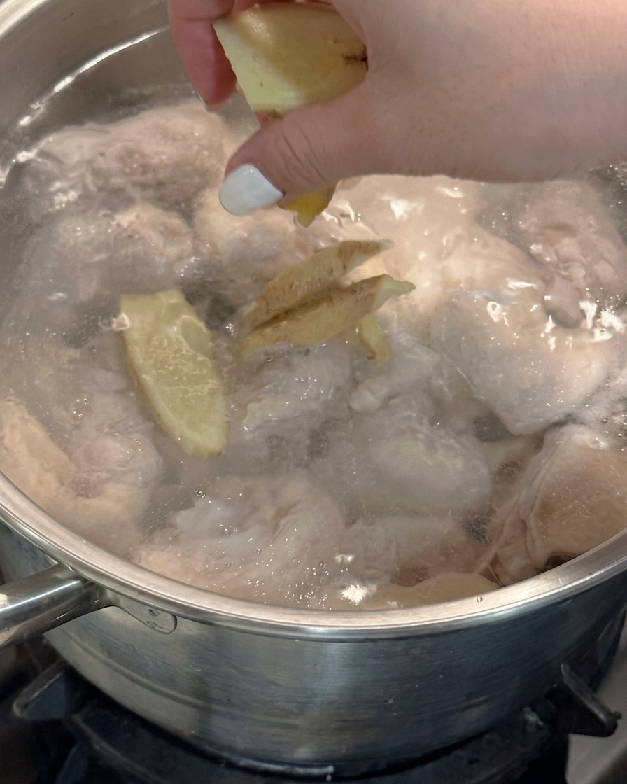 A hand dropping fresh ginger slices into a pot of boiling water with chicken.