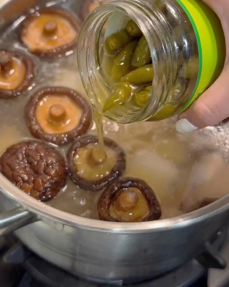 Pickled peeled chili pepper juice being poured from a glass jar into a pot of simmering soup with shiitake mushrooms.