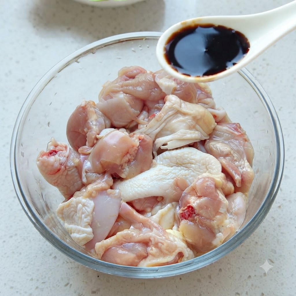 A spoonful of dark soy sauce being added to a glass bowl filled with raw chicken pieces.