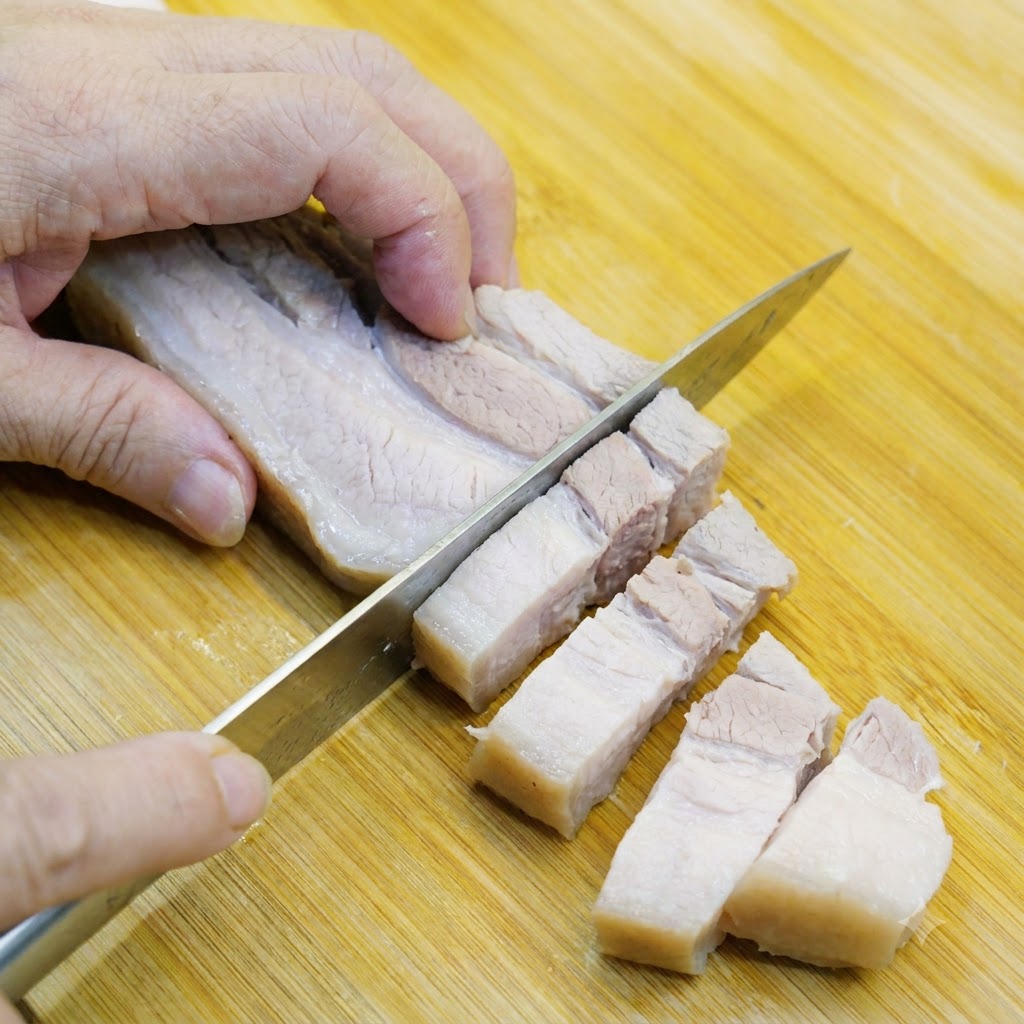 Blanched pork belly being sliced into bite-sized pieces on a wooden cutting board.