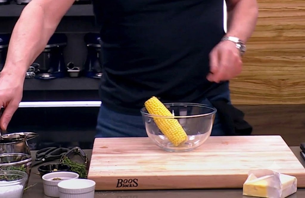 An ear of raw corn sitting in a glass bowl, ready to be seasoned with olive oil, salt, and pepper.