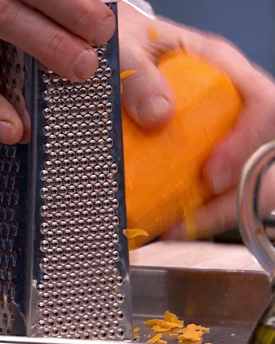 Hands using a metal box grater to shred bright orange butternut squash onto a metal tray.
