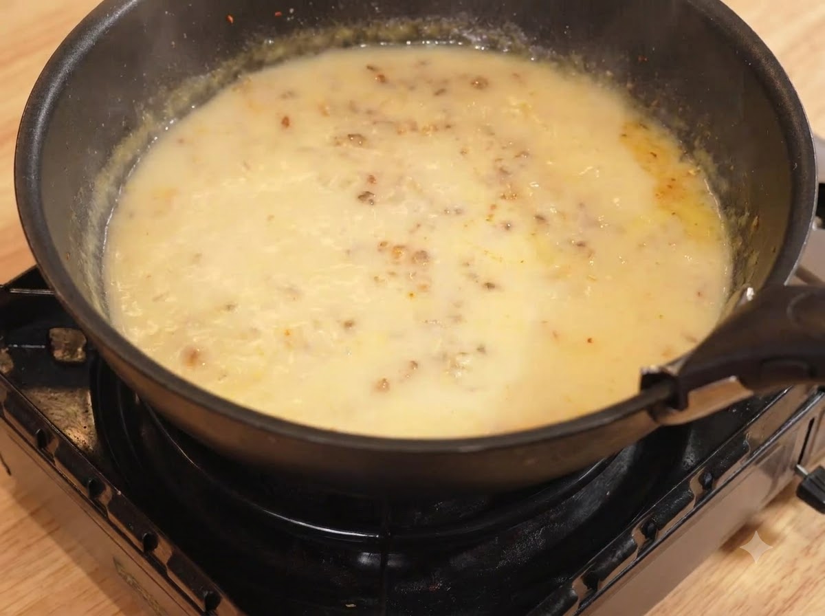 Water and milk being poured into the pan with the curry mixture, simmering over low heat.