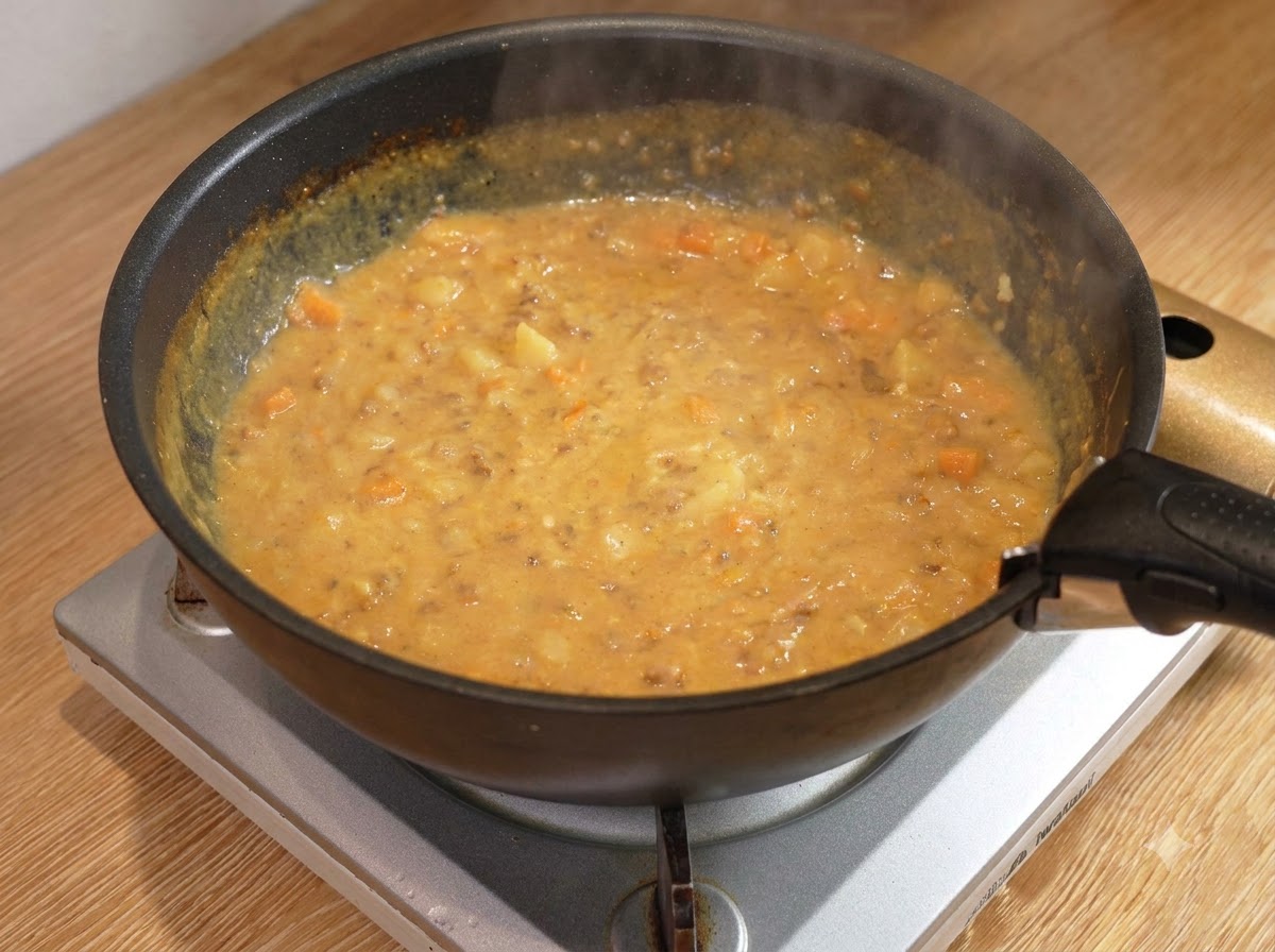 A wooden spoon stirring a thick, bubbling curry meat sauce inside a black pan on a burner.
