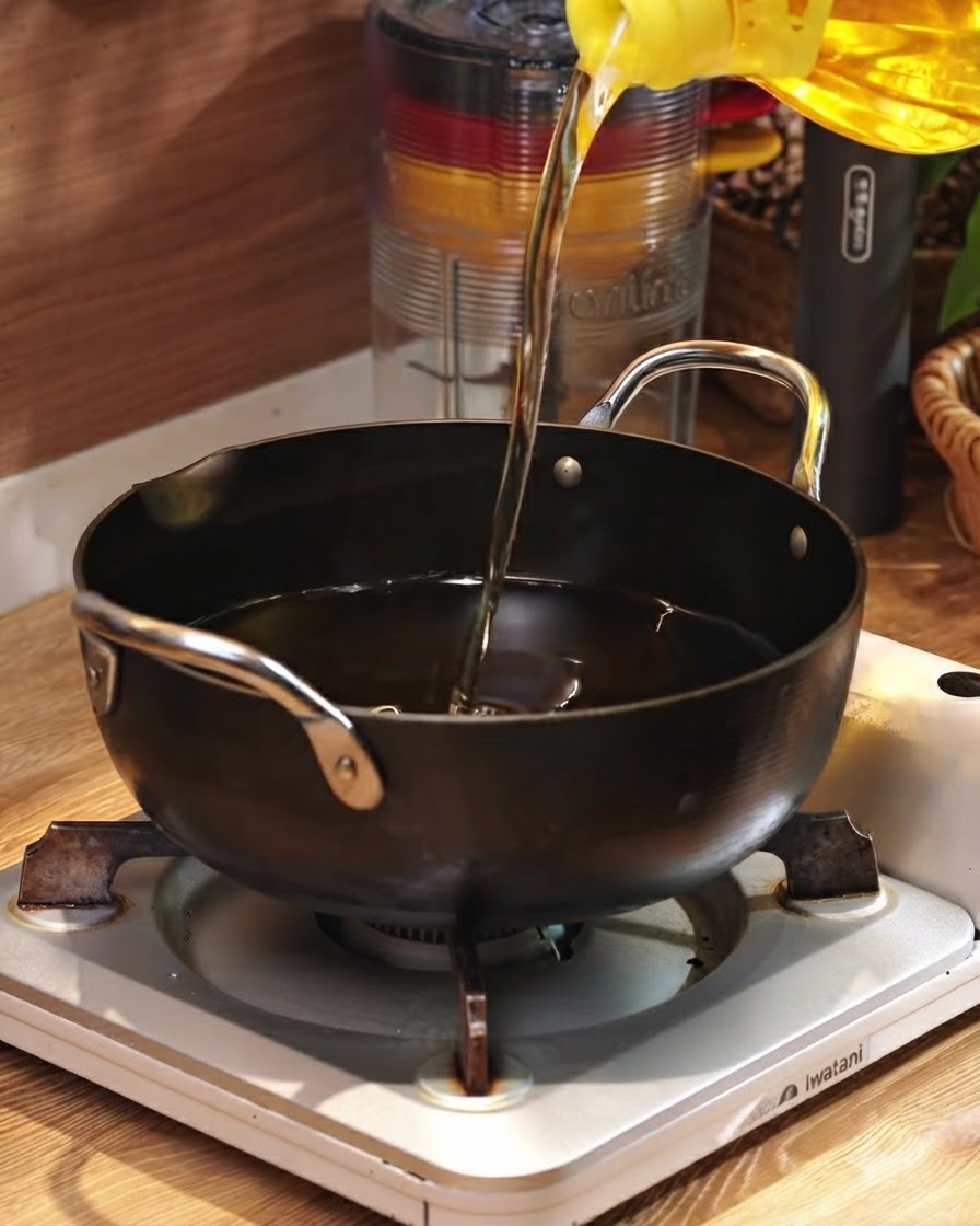 Cooking oil being poured from a yellow container into a black pan resting on a portable burner.