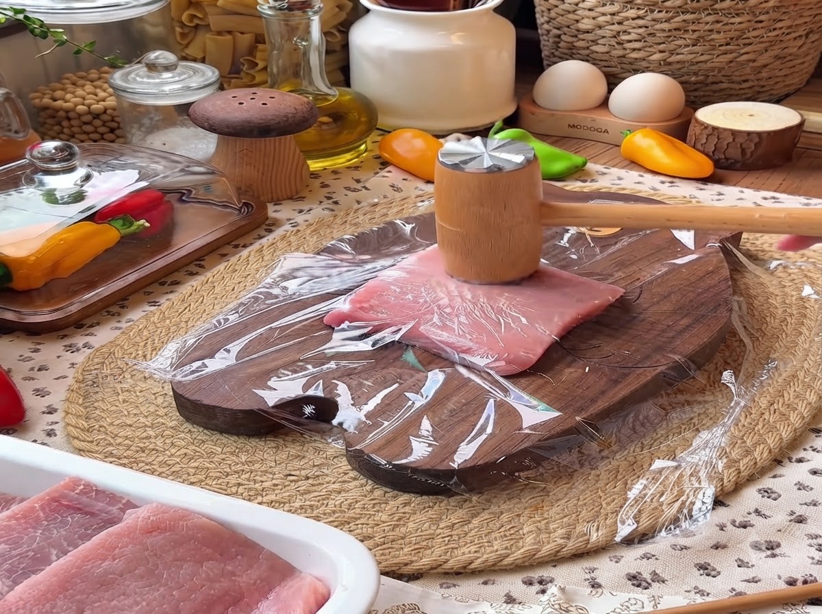 A wooden meat mallet pounding down on a piece of pork tenderloin covered with plastic wrap on a wooden cutting board.