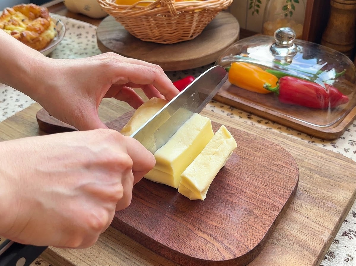 A pair of hands using a knife to slice a block of mozzarella cheese into small rectangular pieces on a wooden cutting board.