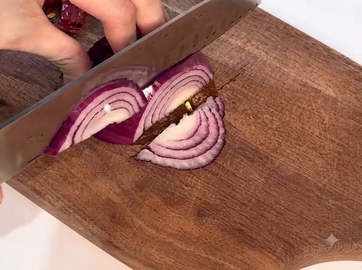 A red onion being carefully sliced on a wooden cutting board with a knife.