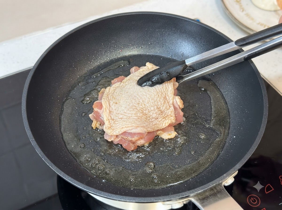 A seasoned chicken thigh frying skin-side down in a hot oiled skillet using tongs.