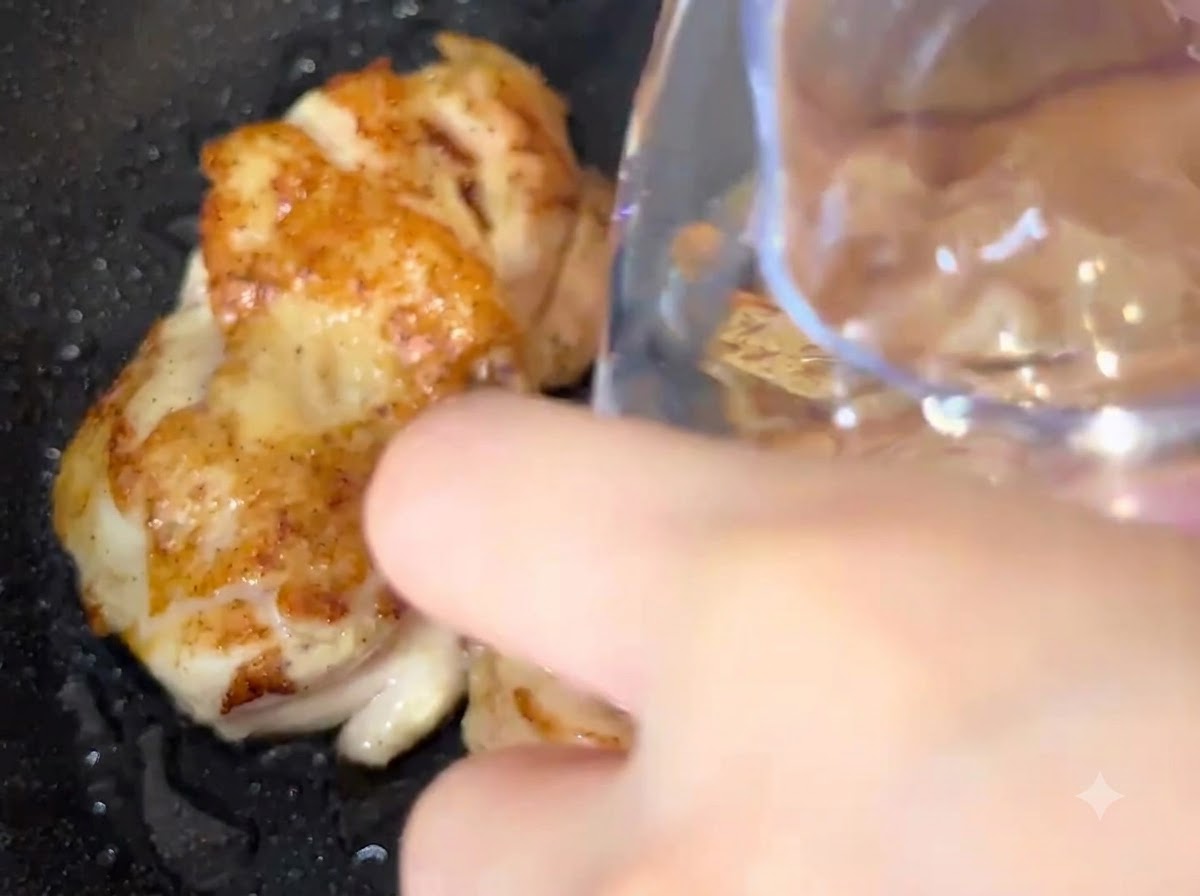 Water being poured from a container directly into a pan with frying chicken pieces.