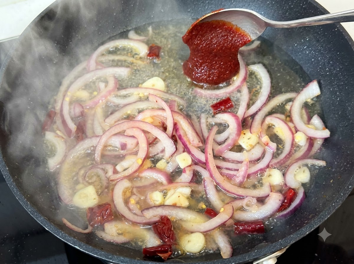 A spoonful of Korean chili paste being added to the simmering pan liquid with onions and spices.