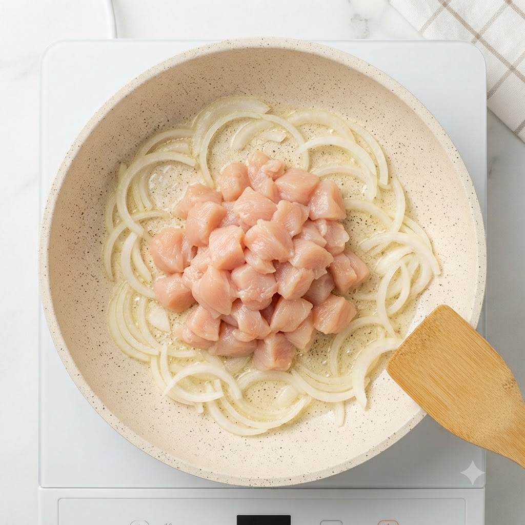 Raw cubed chicken breast piled in the center of a pan surrounded by sautéed onion slices.