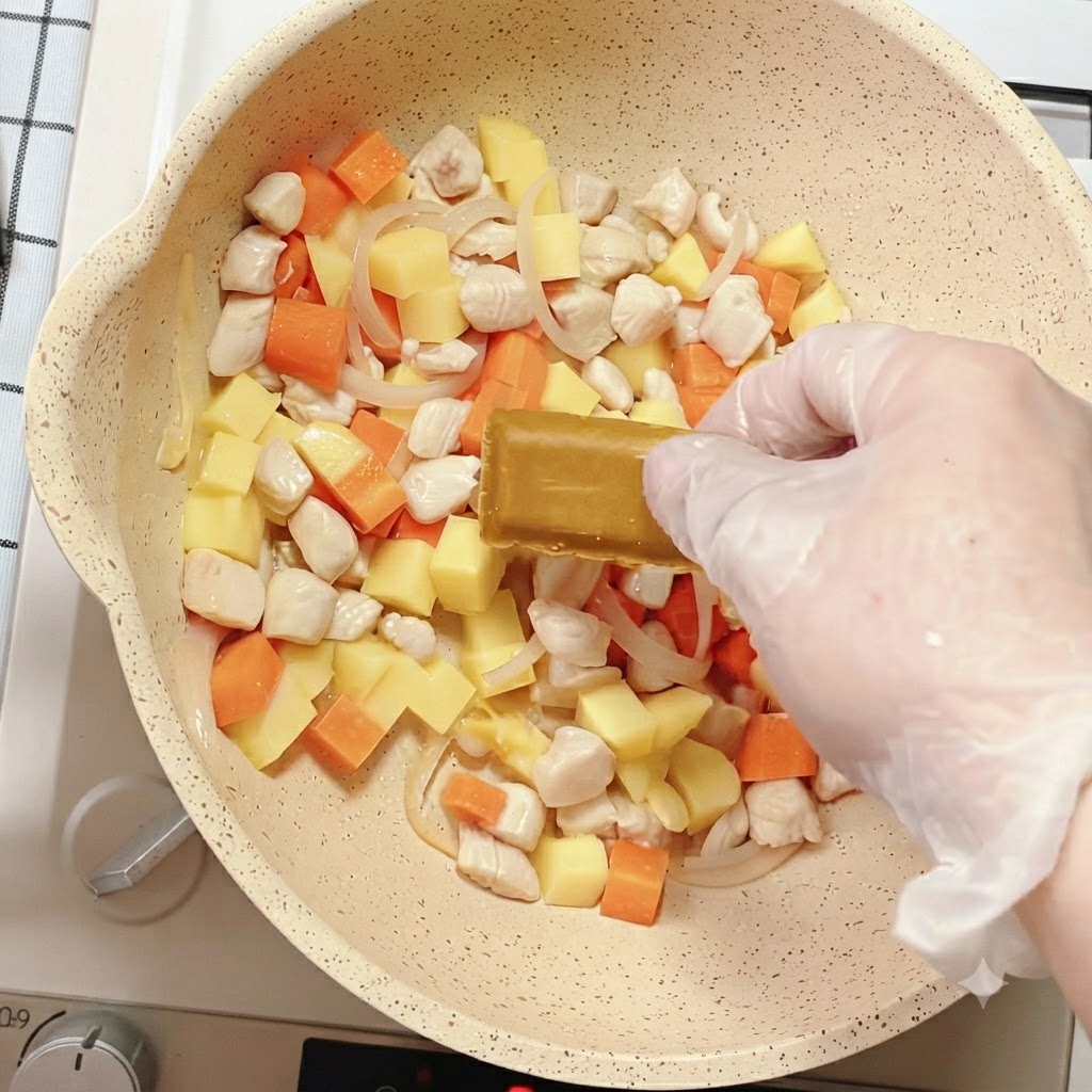 A hand placing a block of curry seasoning into the pan filled with mixed vegetables and chicken.