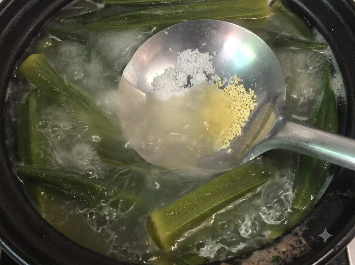 A ladle holding salt and chicken powder over a simmering pot of okra and bone soup.