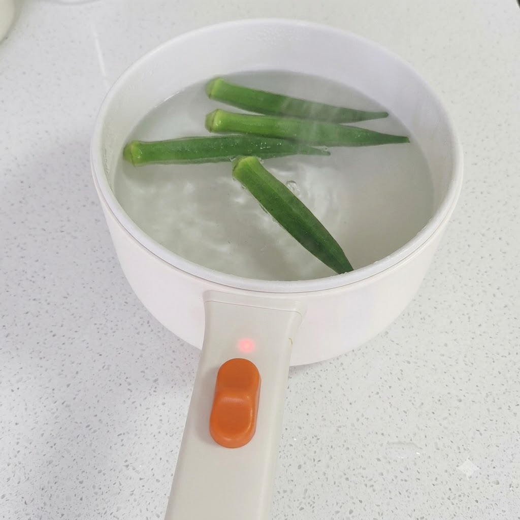 Chopsticks lifting cooked green okra from a pot of simmering water.