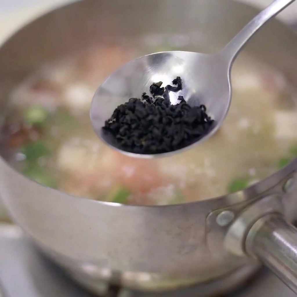 A spoonful of dried seaweed flakes being dropped into a simmering stainless steel pot of soup.