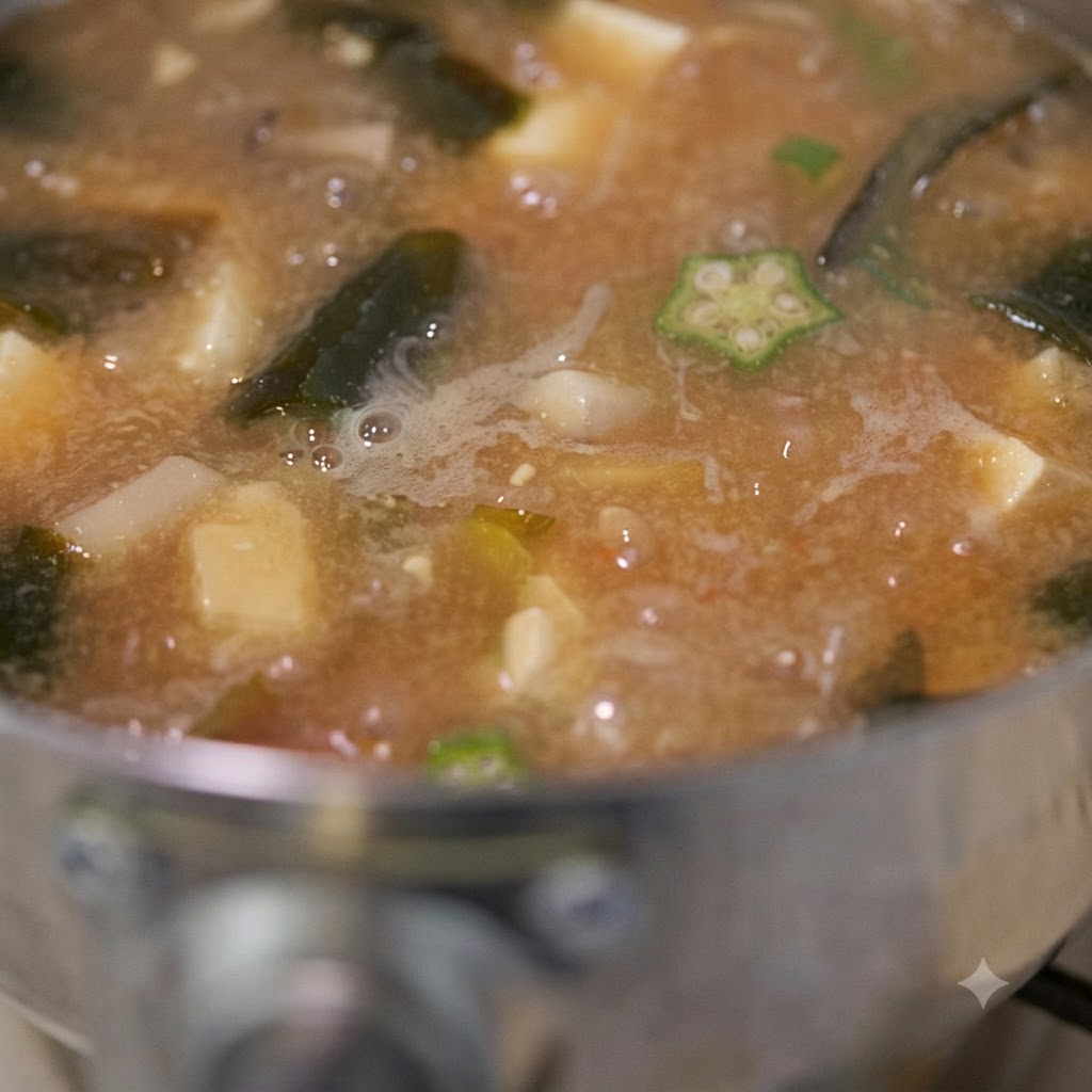 A close-up shot of okra miso soup simmering with visible pieces of okra, tofu, and mushrooms as it is stirred.