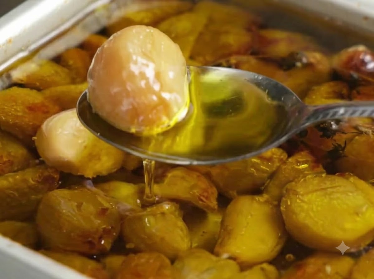 A spoon scooping a golden roasted garlic clove from a baking pan filled with olive oil and garlic cloves.