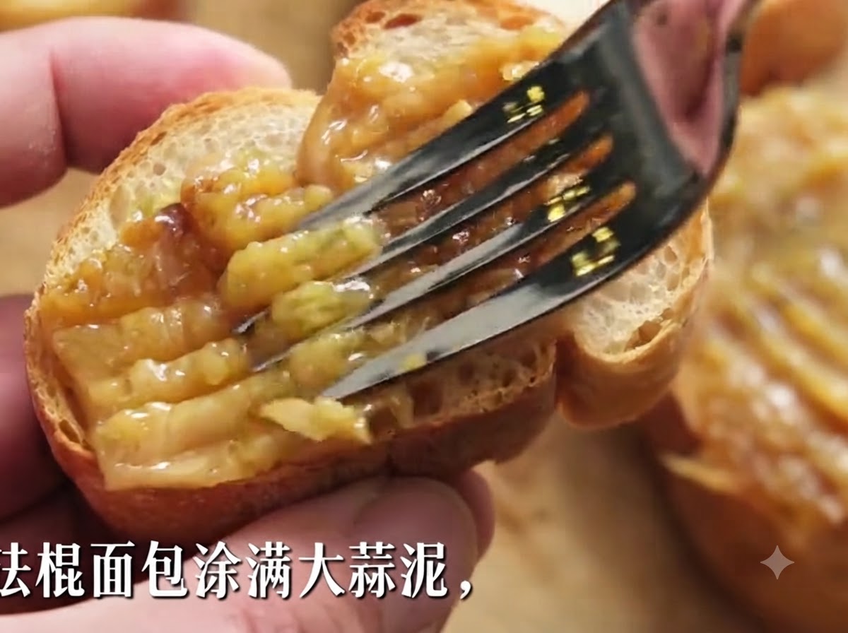 A fork spreading thick roasted garlic paste onto a slice of crispy baguette.