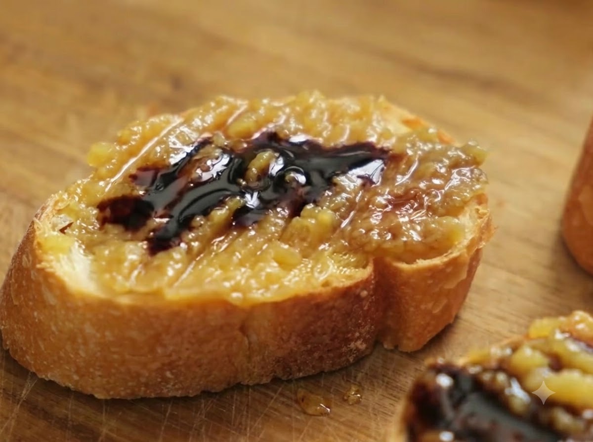 A close-up of a baguette slice topped with garlic confit paste and balsamic vinegar, with coarse sea salt being sprinkled over it.