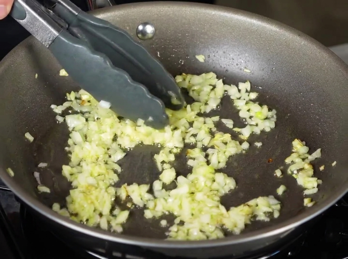 Boiled tagliatelle pasta being placed using tongs into a frying pan with chopped sautéed onions.