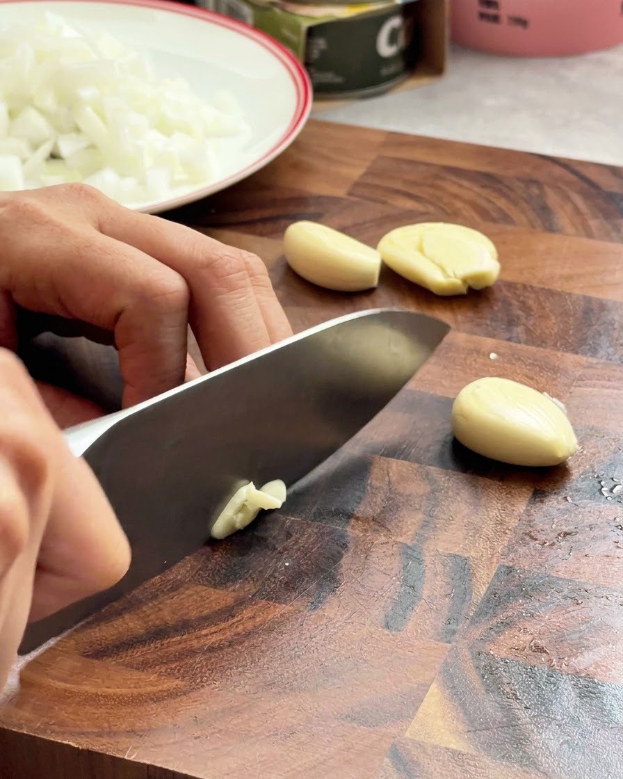 Garlic cloves being minced with a chef knife on a wooden surface.