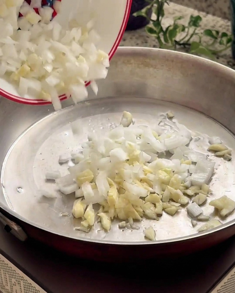 Minced onions and garlic sliding from a small white bowl into a warm metal pan.