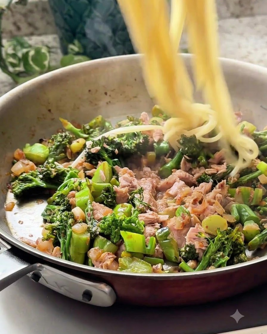 Cooked spaghetti strands being tossed into the pan with the tuna and vegetable mixture.