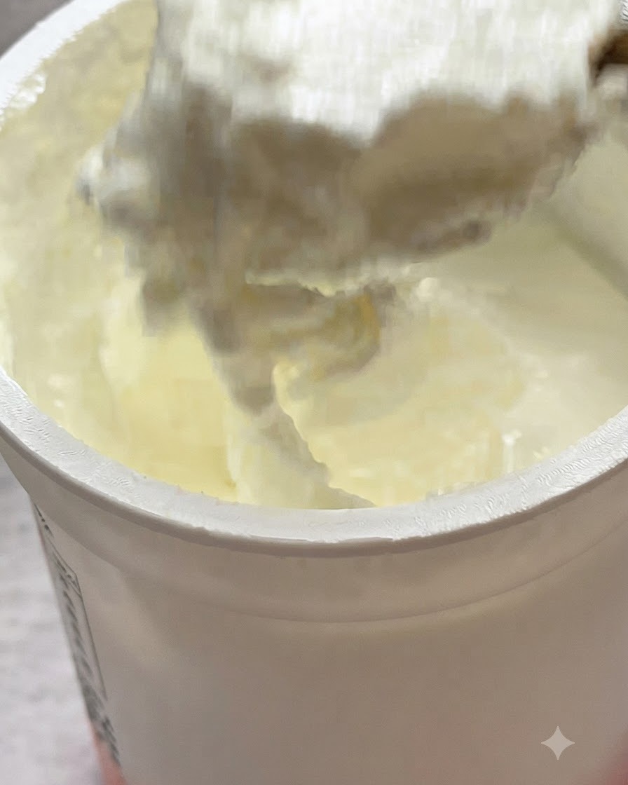 A spoonful of unsweetened yogurt being added on top of pasta in a pan.