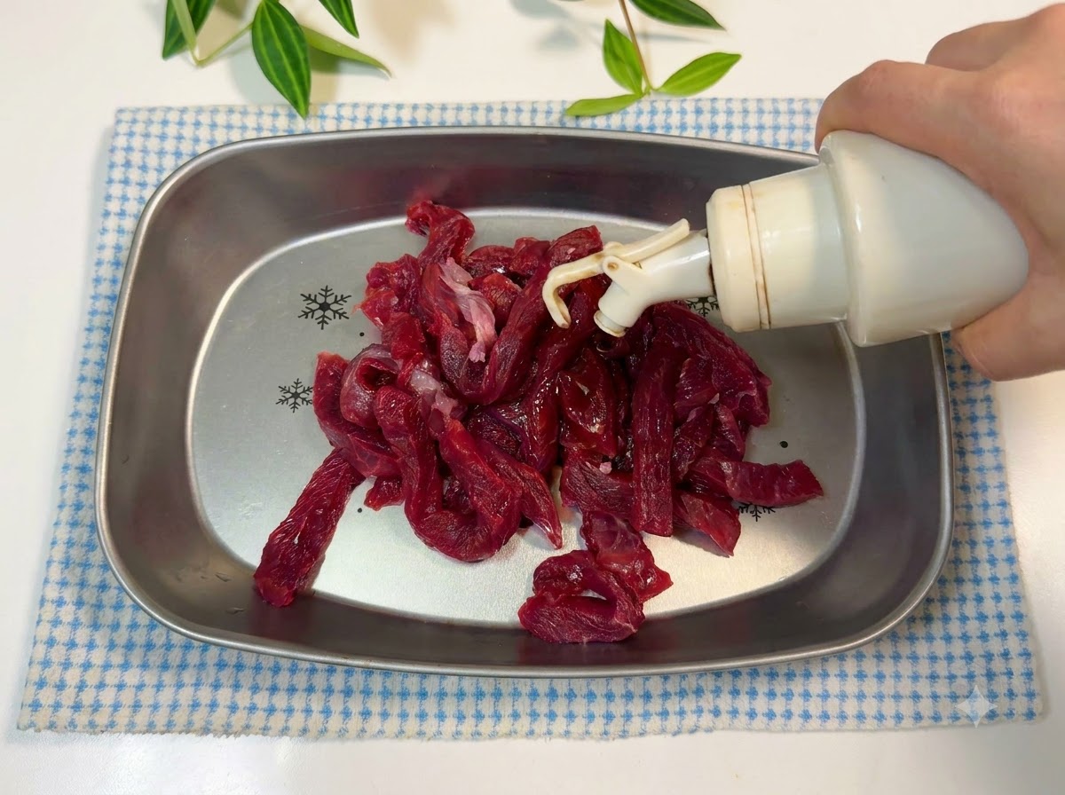 Beef strips in a metal tray being seasoned with oil and soy sauce.