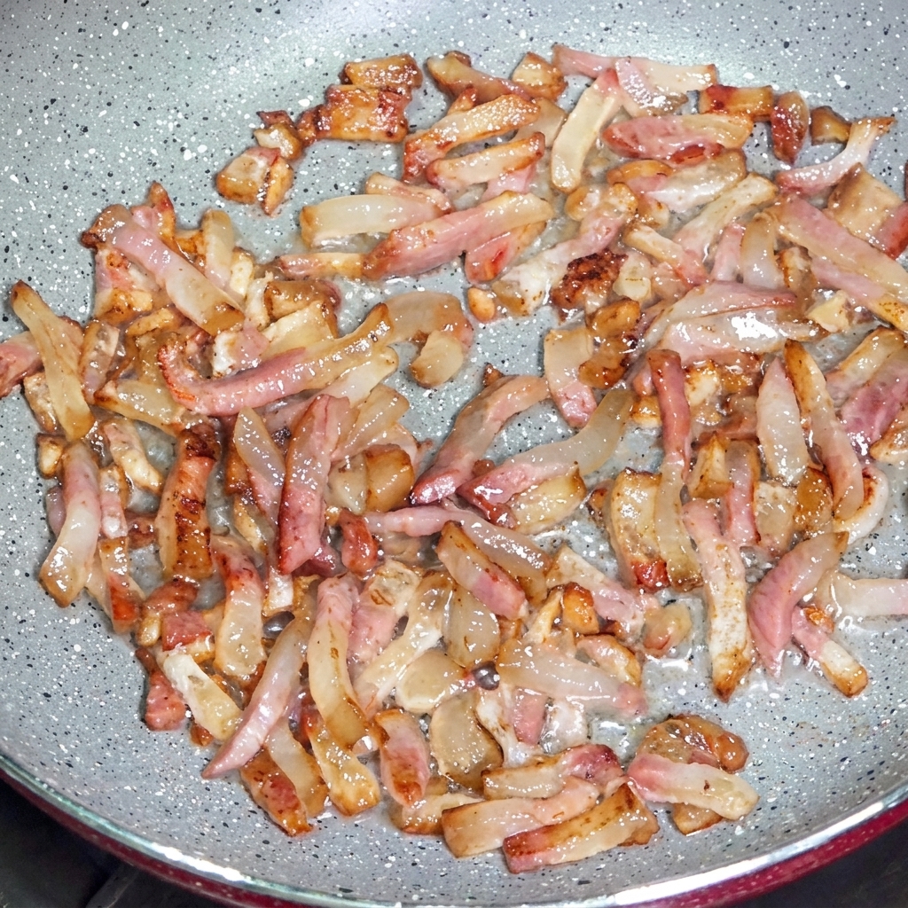 Strips of guanciale frying in a pan, releasing clear fats and developing crispy browned edges.