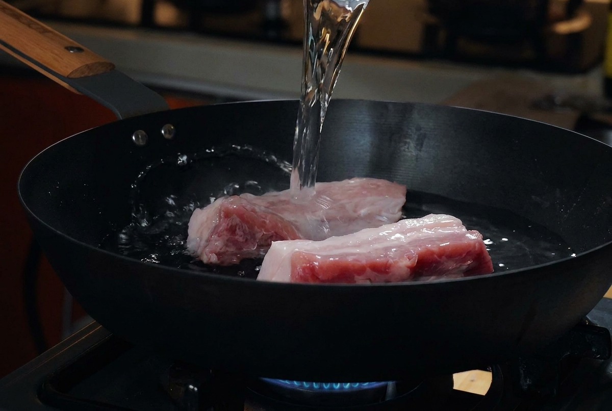Thick cuts of raw pork belly resting in a cold black pan while water is being poured over them from a measuring cup.