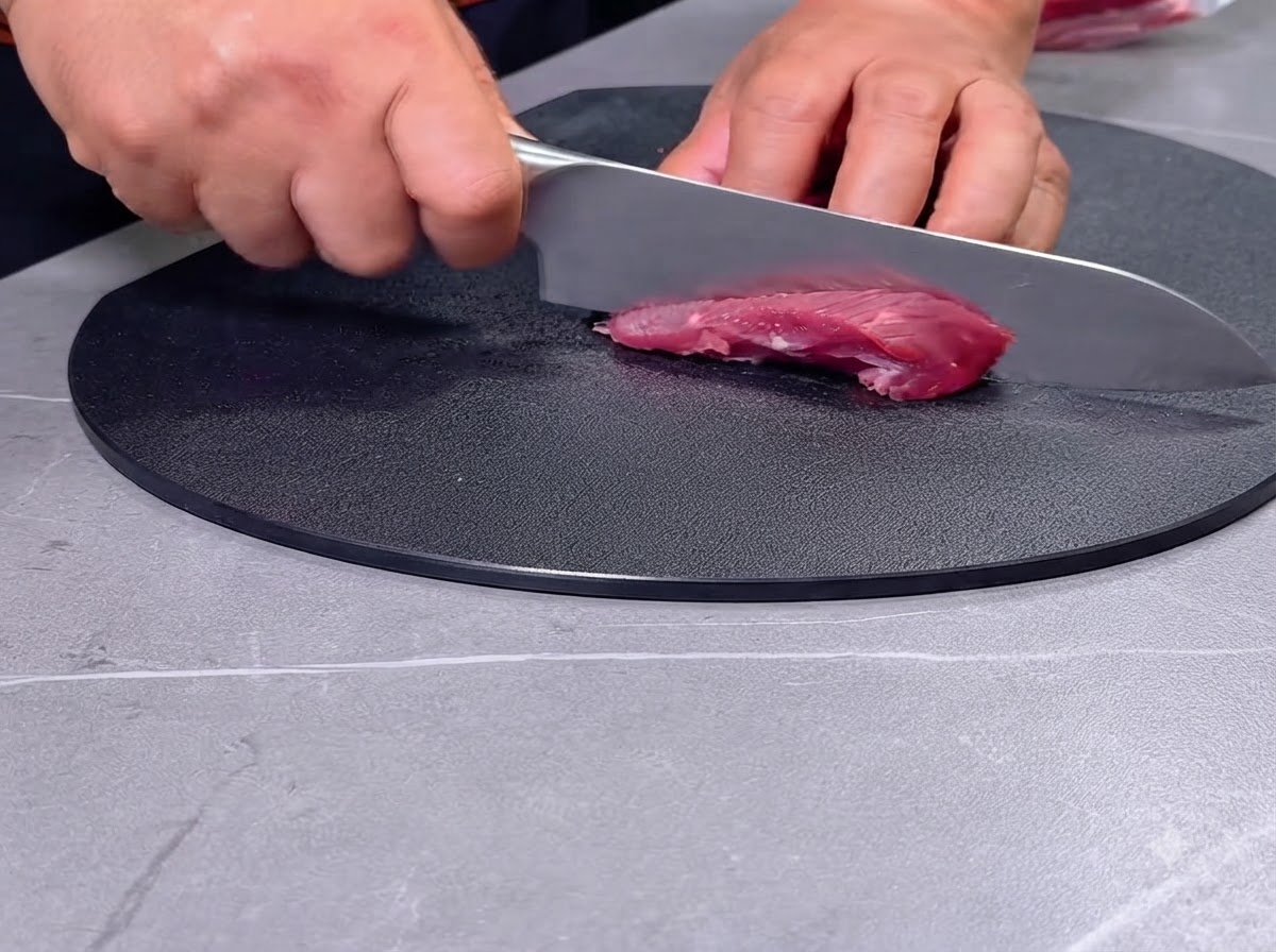 A person slicing raw red meat into chunks with a knife on a black cutting board.